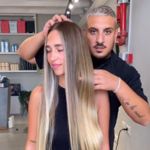 שלומי עומד מאחורי לקוחה עם שיער ארוך ובהיר, ומסדר לה את השיער